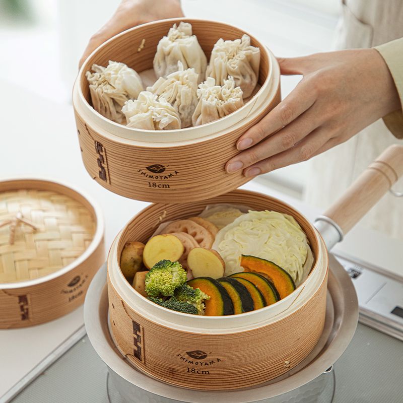 Hands lifting a bamboo steamer tier of dumplings above a tier of steamed seasonal vegetables